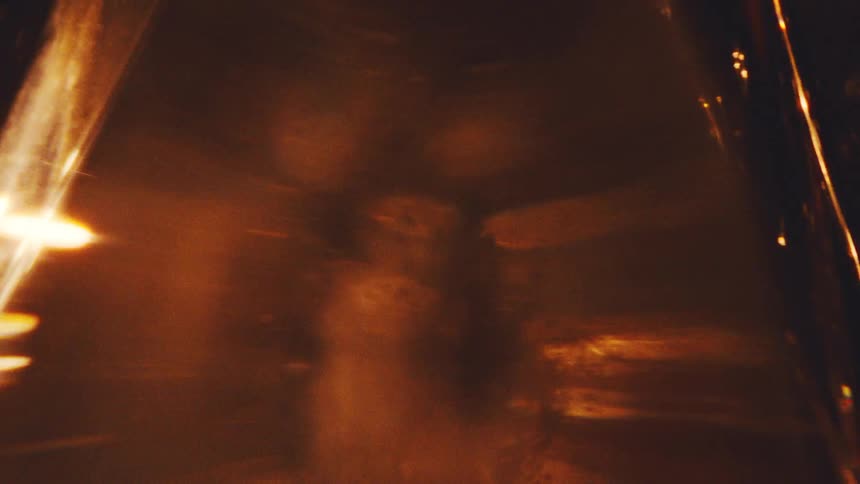 A close view shows warm golden light reflecting and shimmering through a textured glass surface against a dark background creating abstract shapes and bright flares