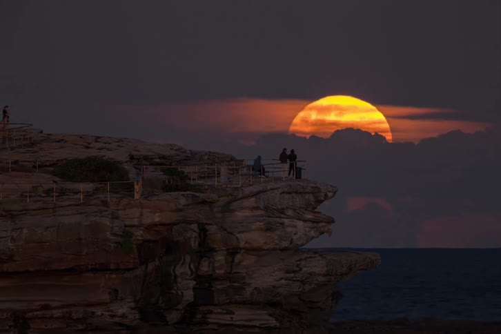 Full moon rises on the horizon behind rocky seaside formation at dusk