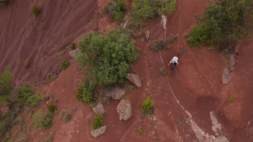 A mountain biker navigates a narrow rocky path down a steep hillside characterized by vibrant red earththe aerial perspective captures the rider maneuvering around sparse green bushes and large stones as they descend the rugged terrain