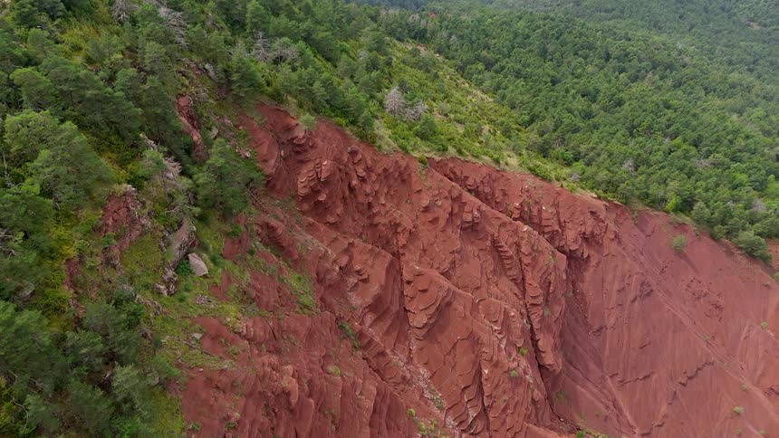 A massive red rock canyon with jagged ridges and steep slopes surrounded by a dense green pine forest