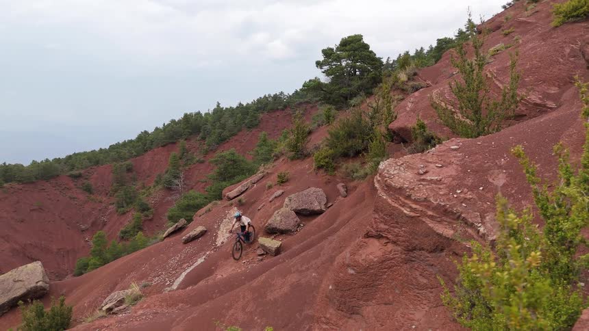 A person rides a mountain bike down a steep and rocky red dirt slope on a hillside with green trees and bushes under a cloudy sky