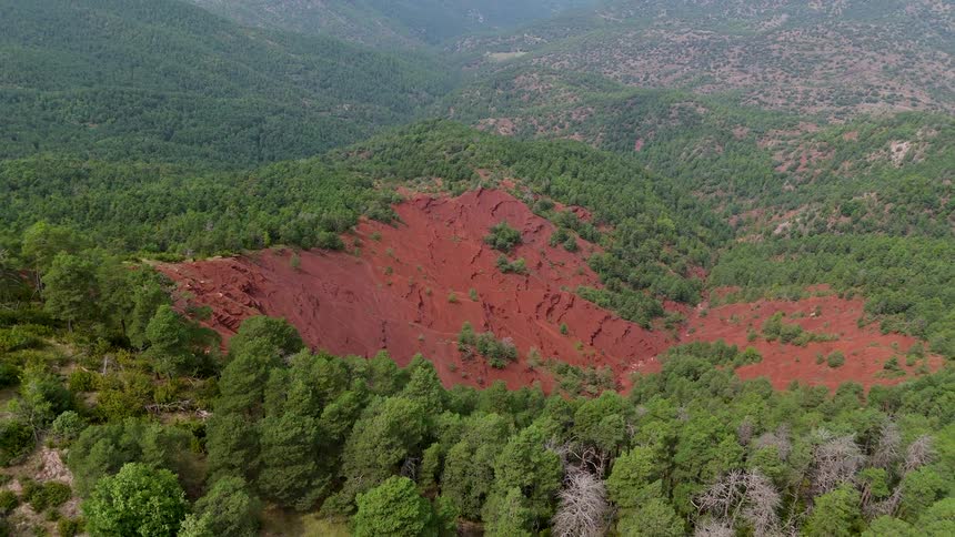 An aerial view drifts over green forested hills and a broad scar of red earth sliding past treetops as the reddish slope falls away and the hazy mountain ridges stretch across the background