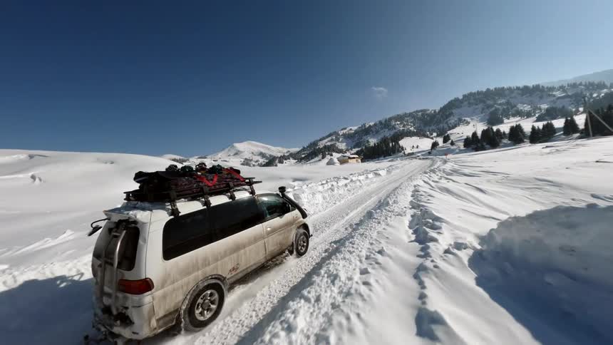 Vehicles travel over a snowy road through the rural countryside near forested mountains on a sunny winter day