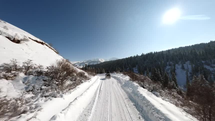 A silver suv and a black truck driving along a narrow snow covered road winding through a steep mountain valley under a bright sun with dense evergreen forests
