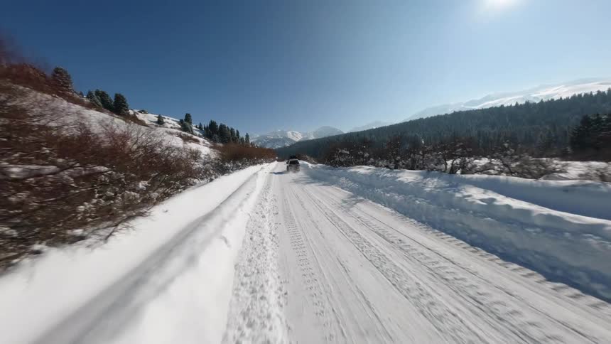 A drone flies over a snowy mountain road as several vehicles drive through a winter landscape with pine trees and high peaks under a clear blue sky