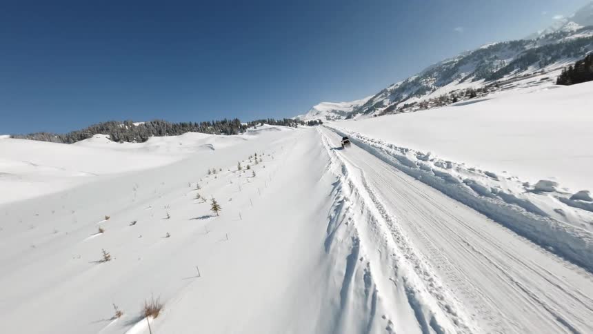 A white off road van with a roof rack drives up a steep snow covered mountain path surrounded by pine trees and distant alpine peaks under a clear blue sky with a bright sun