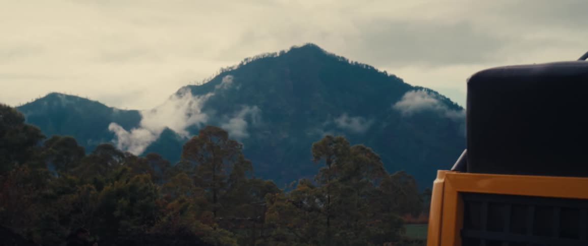 Mistcovered mountains in the background with trees in the foreground and part of a yellow vehicle with a black tank on the right