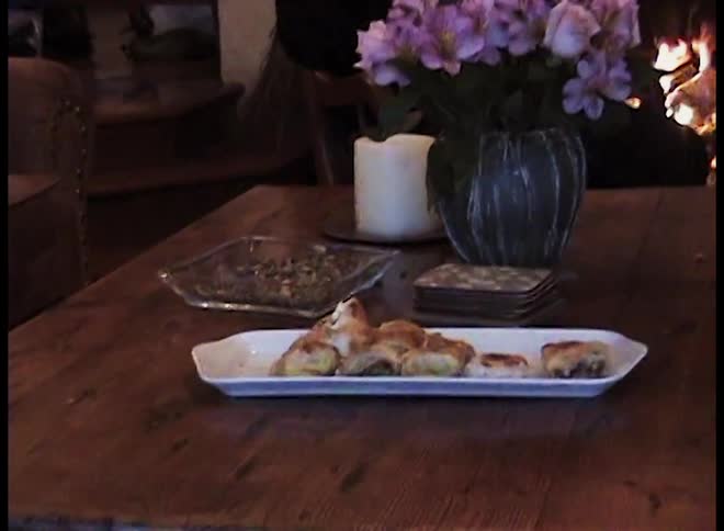 A wooden table with several small pastries arranged on a rectangular white platenearby there is a large white candle a glass dish with snacks and a vase of purple flowers