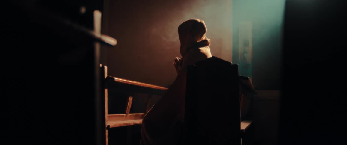 A person draped in a heavy cloak sits in a dark wooden pew hands clasped in prayer as soft light and subtle smoke filter through the quiet dim atmosphere of a church