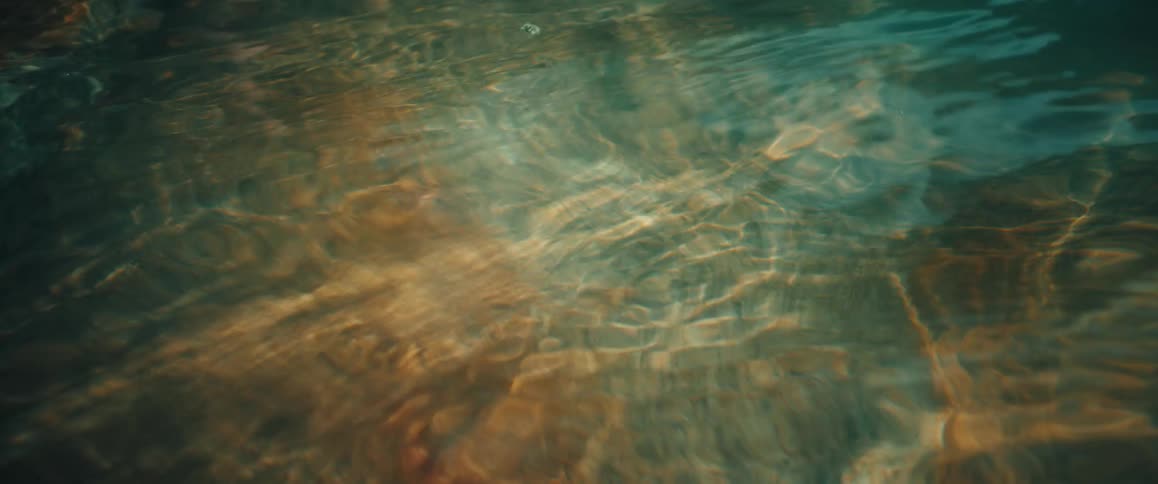 The shimmering surface of clear shallow water as sunlight creates dancing patterns on the sandy floor belowgentle ripples move across the frame distorting the golden and teal hues of the water in a calm rhythmic motion