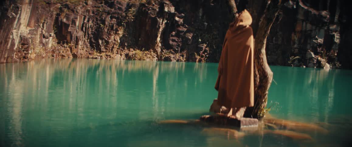 A mysterious figure draped in a long brown cloak stands motionless on a tiny stone island beside a weathered tree overlooking the still turquoise waters of a secluded quarry as sunlight illuminates the jagged rock face