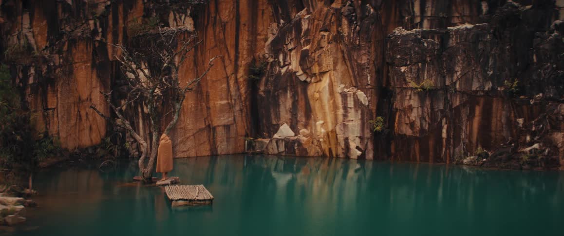 Steep mountain cliff overlooks a woman wearing a hooded cloak standing on a rock near her raft in the water next to a tree