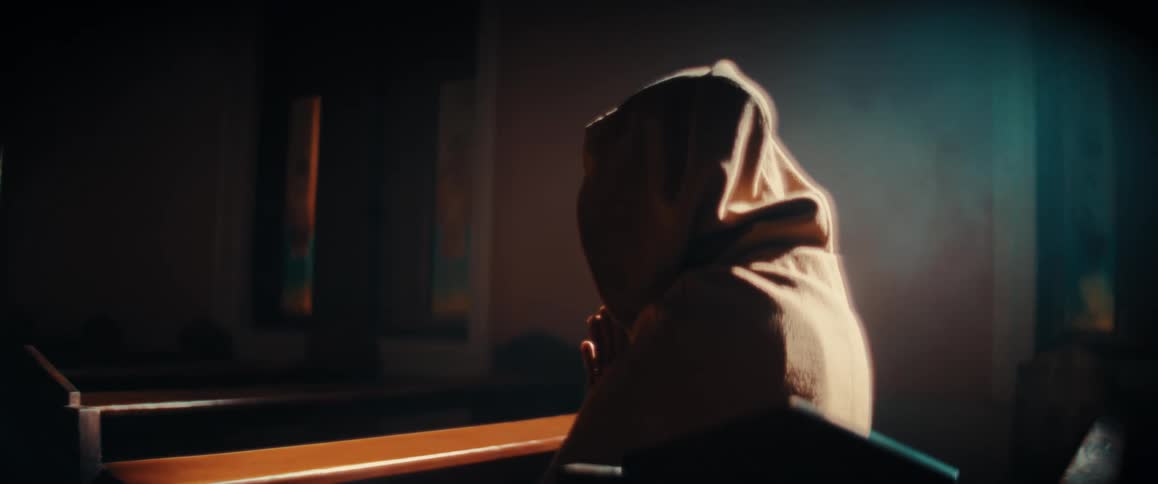 Female person wearing a gown with a hoodie covering her head praying with her hands clasped siting on a wooden bench inside of a church during day time