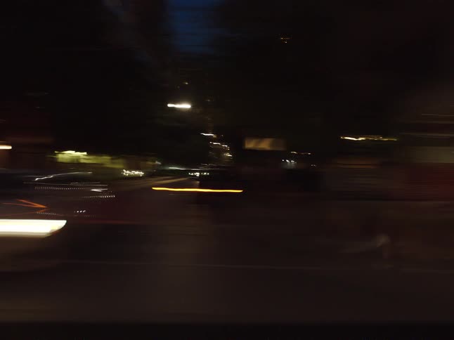 A blurry night scene from a moving vehicle as it passes through an urban environment with glowing orange street lights and dark building structures
