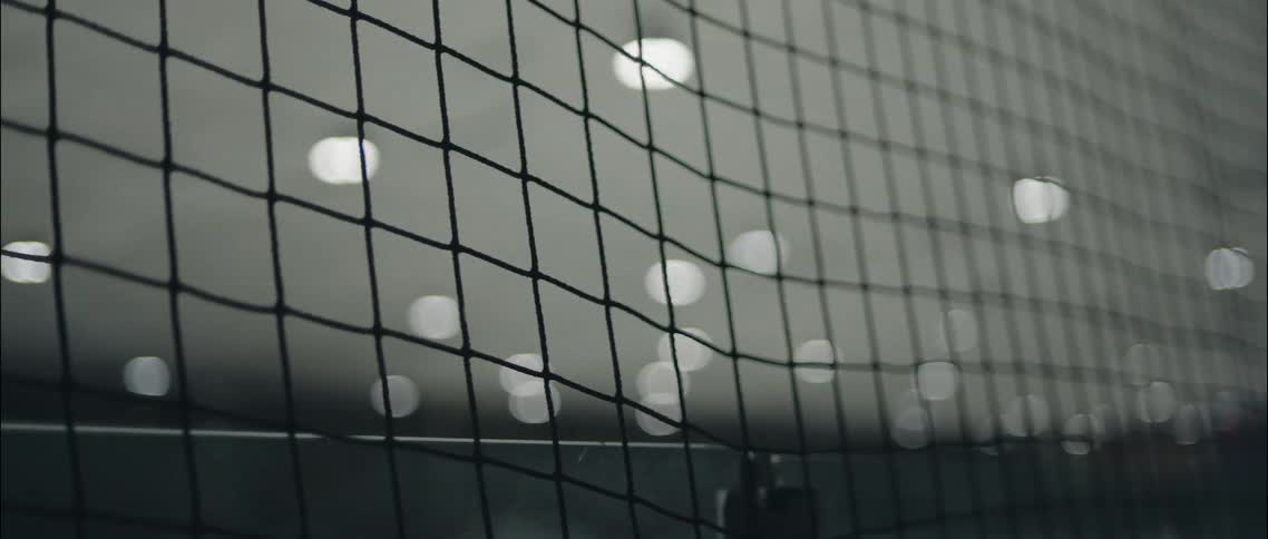 A closeup shot focuses on the intricate knots of a black safety net in a sports arena while overhead stadium lights create a soft blurred bokeh effect in the dark background