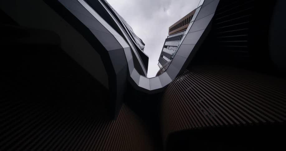 The video presents a lowangle perspective looking up at modern architectural structures with sharp angular designs and textured surfaces a cloudy sky is visible through the narrow gap between the towering buildings creating a sense of depth and scale