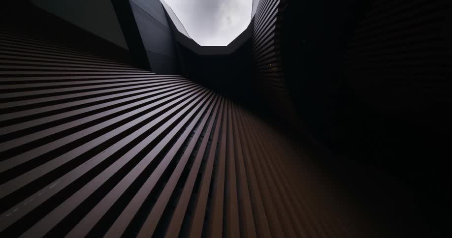 An upward view of a modern architectural structure featuring dark parallel slats that converge toward a bright opening in the sky