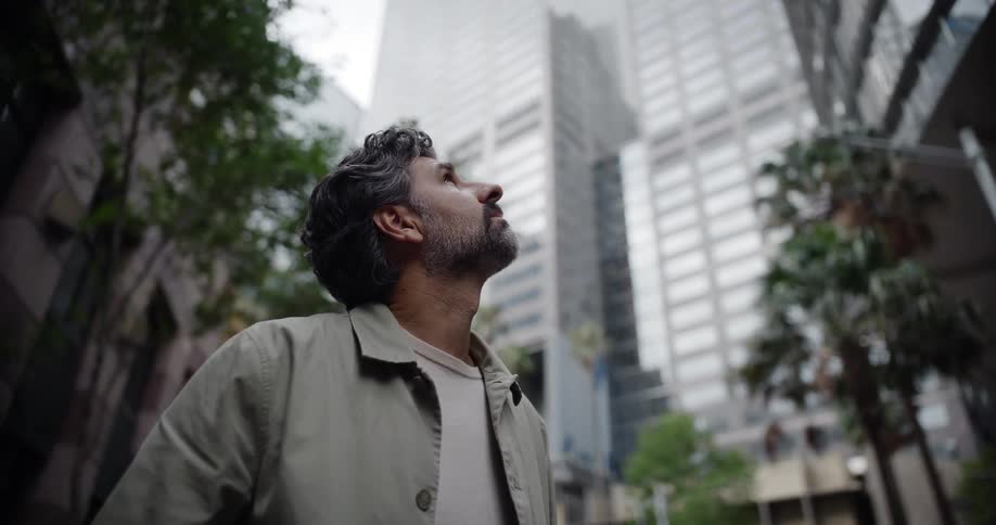 Man looking upward between tall buildings in an urban area with trees around