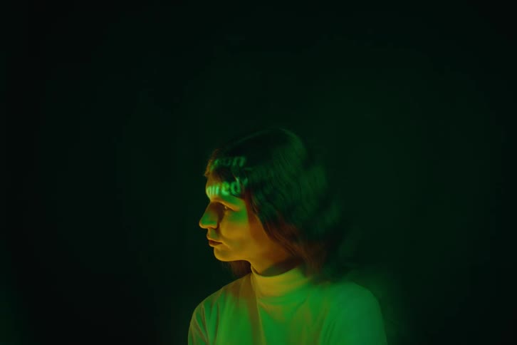 A young woman in profile illuminated by green and yellow light against a dark background various words and patterns are projected directly onto her face and hair creating a flickering cinematic effect as she slowly shifts her gaze
