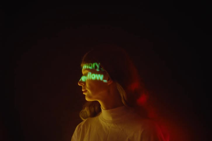 Androgynous adult person wearing a white blouse with green text projected over their face in a room with red lights indoors