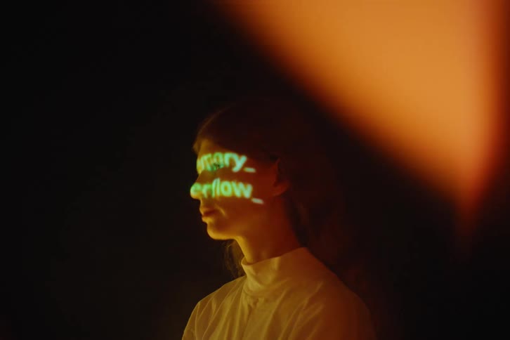 A woman in profile is illuminated by warm light as green projected text patterns glow across her face against a dark background