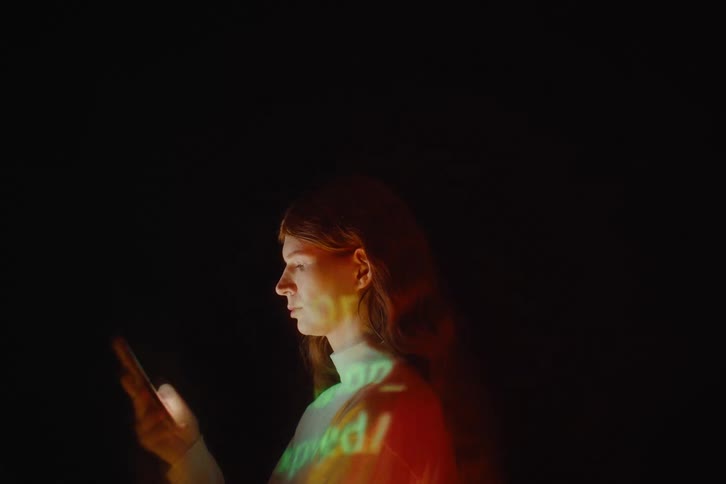 A woman in profile looking at her smartphone while colorful light patterns and text are projected across her face and body the atmospheric lighting shifts between dark shadows and bright warm washes of light creating a surreal and techfocused aestheti