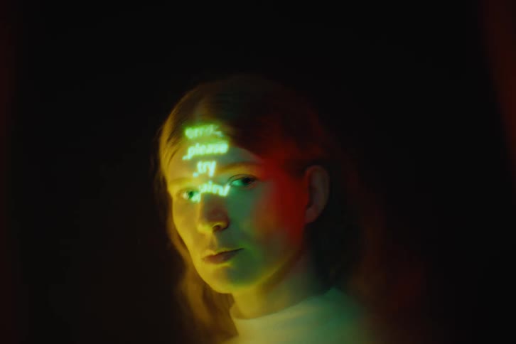 Womans face lit by green projection with text across her forehead in a dark setting