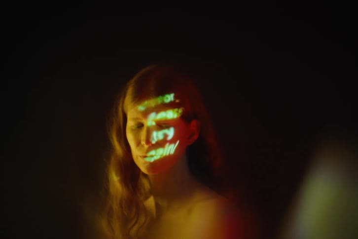 A woman from the upper arms up is illuminated by warm light as green projected text patterns glow across her face against a dark background