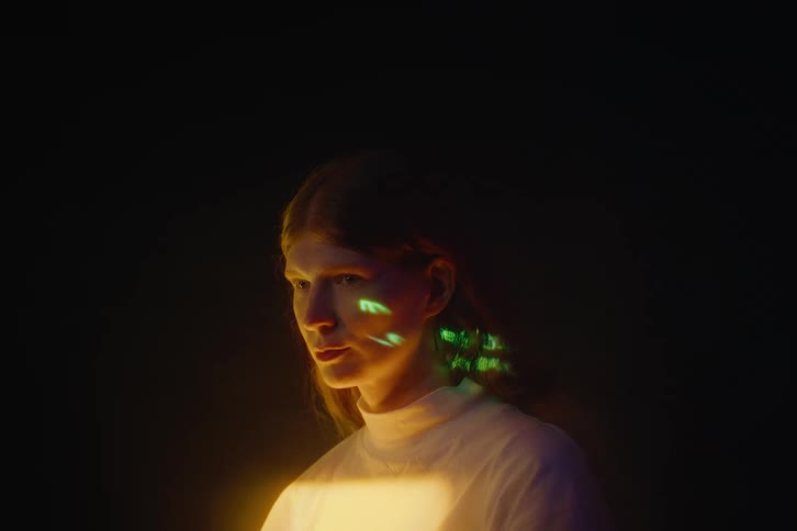 A woman with red hair is lit by vibrant purple green text and warm yellow light against a dark background creating a dramatic futuristic portrait