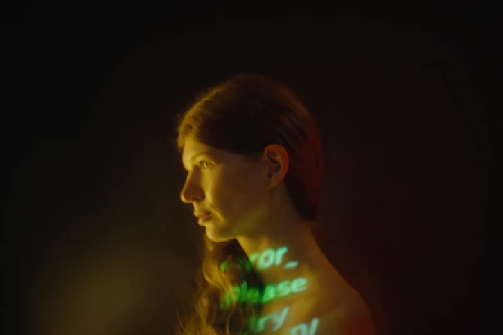 A person with long hair turns their head with green digital text projected onto their shoulder and back on a dark background