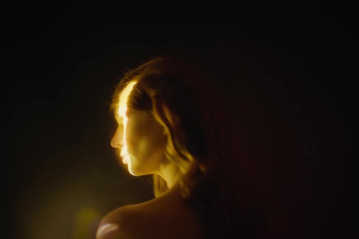 A woman stands in profile against a dark background illuminated by a narrow bright beam of warm light across her faceshe slowly turns her head away from the camera allowing the light to trail over her long wavy hair as vibrant red and orange lens f