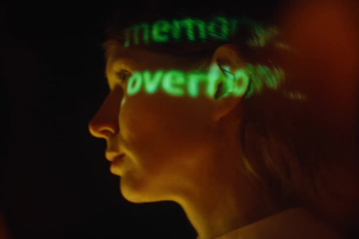A young woman stands in a dark room as a projector screen reflects light on her face and behind her