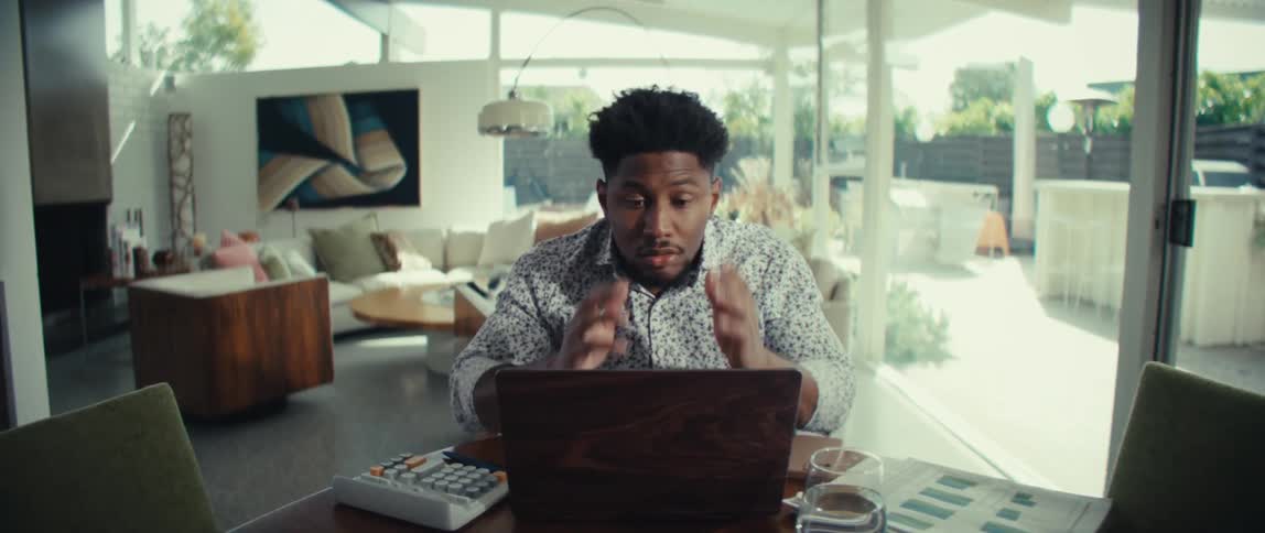A man sits at a table in a bright modern living room gesturing animatedly with raised hands as he reacts to something on his laptop