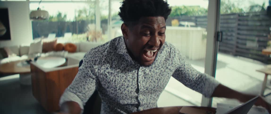 A young man sits at a wooden table and reacts with immense joy while looking at his laptop