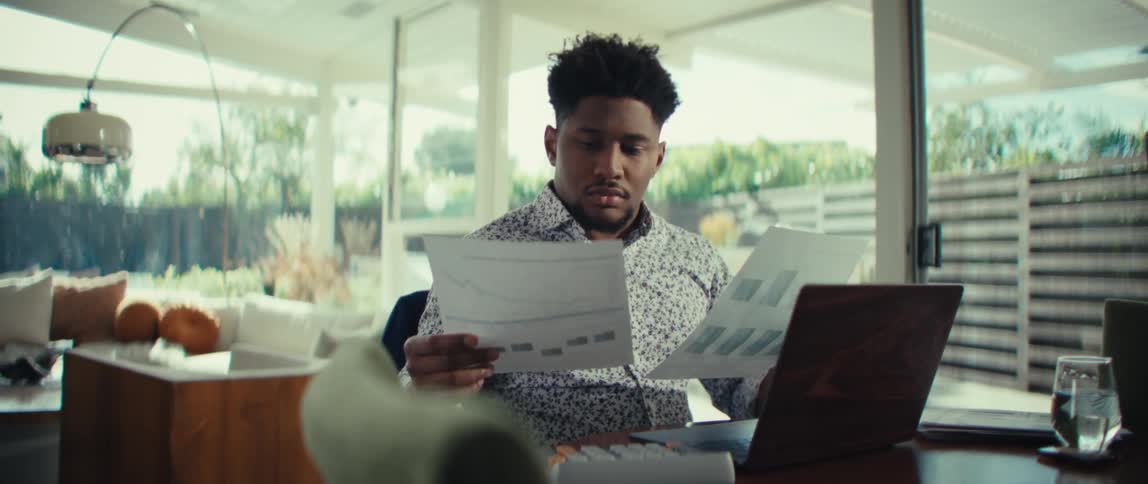 Young black adult man wearing a dress shirt sitting down and reading papers in front of a laptop computer and a calculator over a wooden desk in a living room indoors during day time