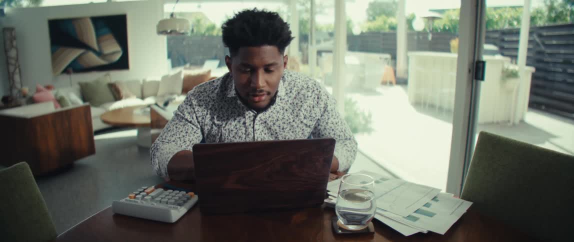 A man in a patterned shirt sits at a dining table focused on his laptop while using a calculator and reviewing financial documentshe works in a bright modern room with large glass doors that look out onto a sunlit patio and backyard