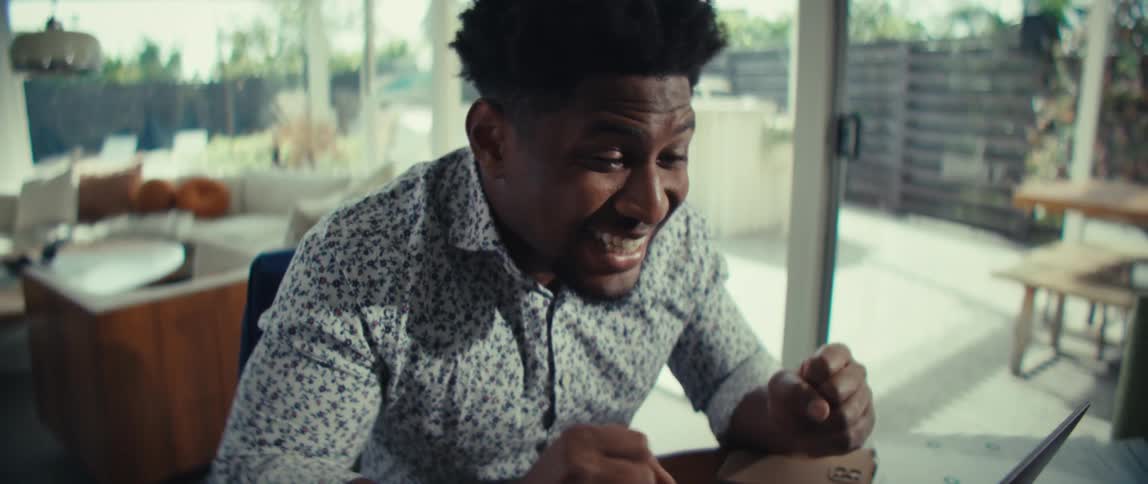 Young adult black man wearing a white dress shirt sitting down in front of a laptop while talking and smiling indoors during day time