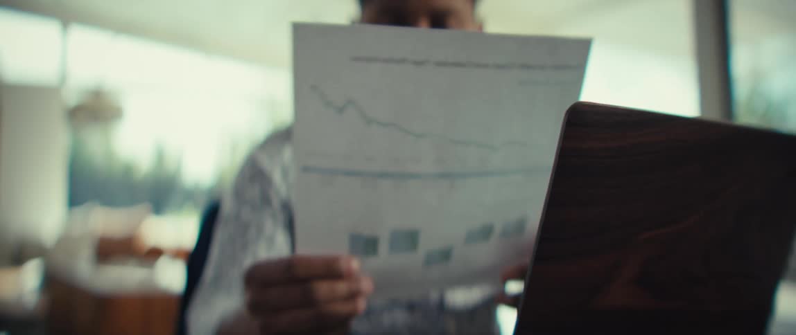 A man in a patterned shirt looks at a document with graphs and charts while sitting next to a laptop