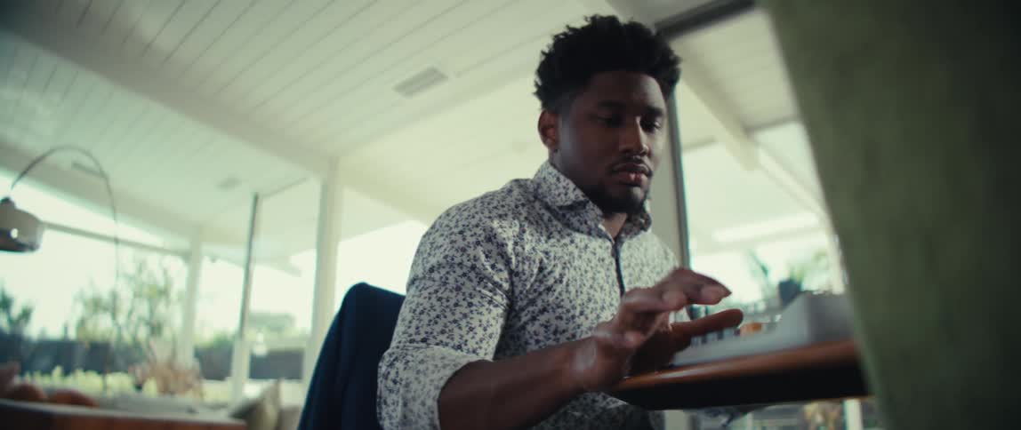 A man with dark hair wearing a patterned shirt sits at a desk and types on a computer keyboard inside a bright room with large windows