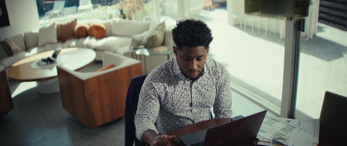 A man sits at a desk in a bright modern living room focused on his laptop with papers spread out beside him