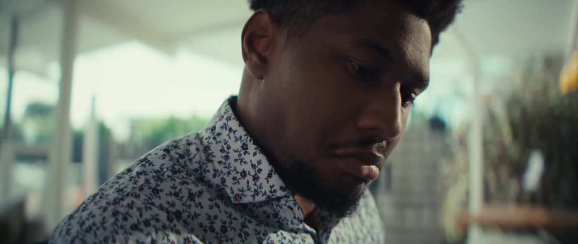 A close view shows the face of a man with dark hair and a beard wearing a floral shirt as he looks down and smiles inside a bright room