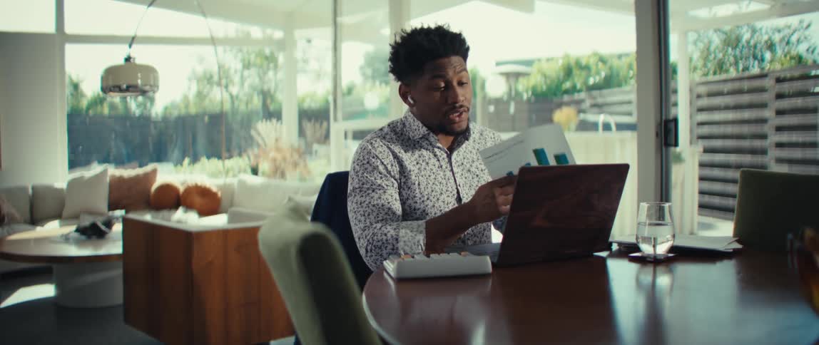 A man is sitting at a wooden table while working on a laptop and wearing wireless earbuds he holds up a printed document featuring blue bar charts and points to the data while speaking as if presenting during a video call