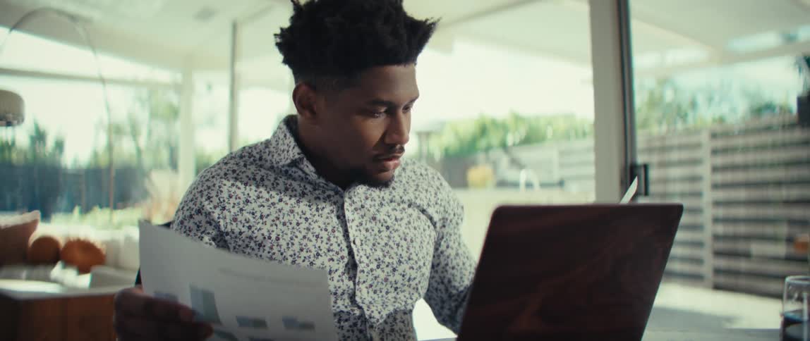 A man sitting at a desk in a bright modern room looking through several printed documents he holds the papers up to examine them closely occasionally glancing toward a laptop screen while appearing focused and thoughtful