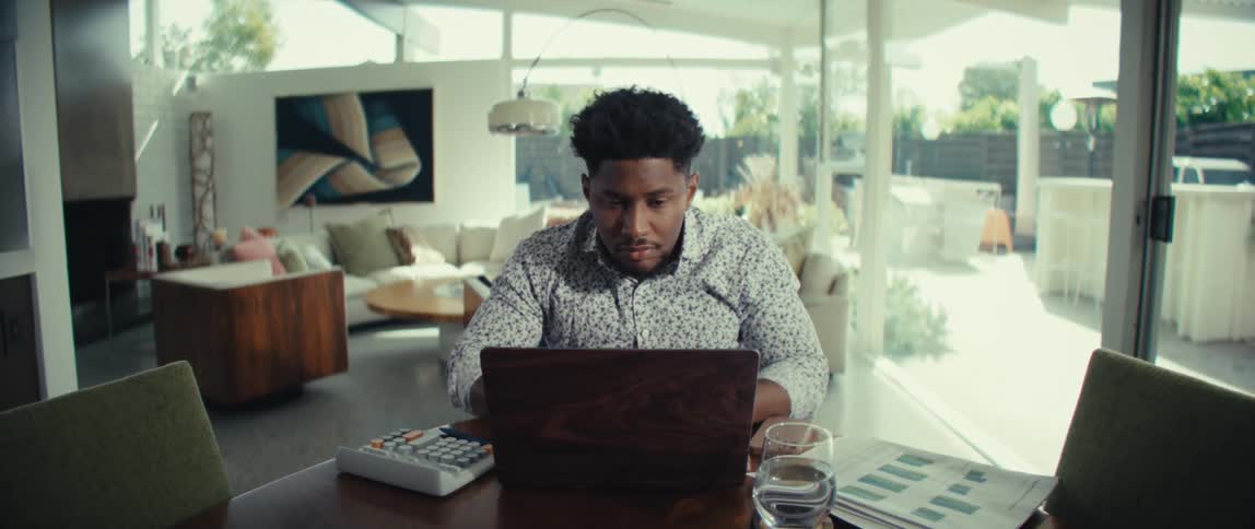 A man wearing a patterned shirt sits at a dining table in a modern home and works on a laptop with a calculator and paperwork nearby as he shows a frustrated expression