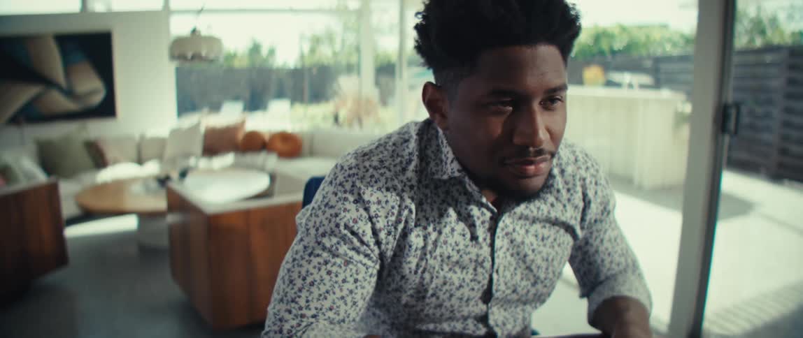 A man wearing a patterned shirt is shown from the chest up working diligently on a woodgrain laptopas the video progresses he pauses his typing to look toward the right of the frame with a slight smile