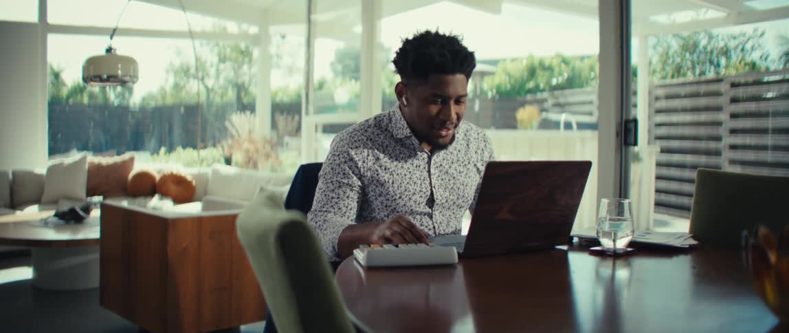 Man gesturing with his hand while using a laptop and calculator at a table in a bright room