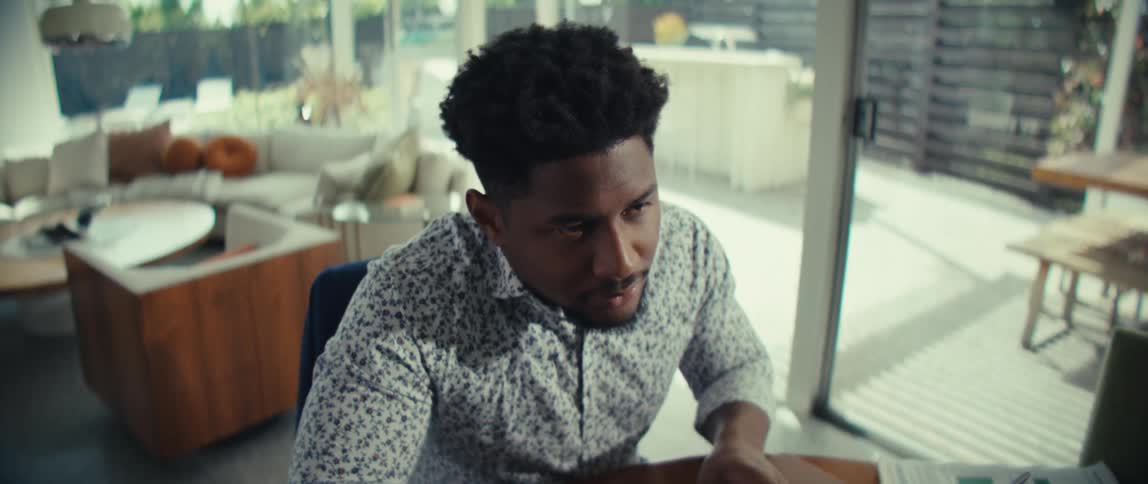 A man in a patterned shirt sits at a round wooden table focused on typing on a laptopas the camera angles shift he pauses his work to look thoughtfully toward a bright window