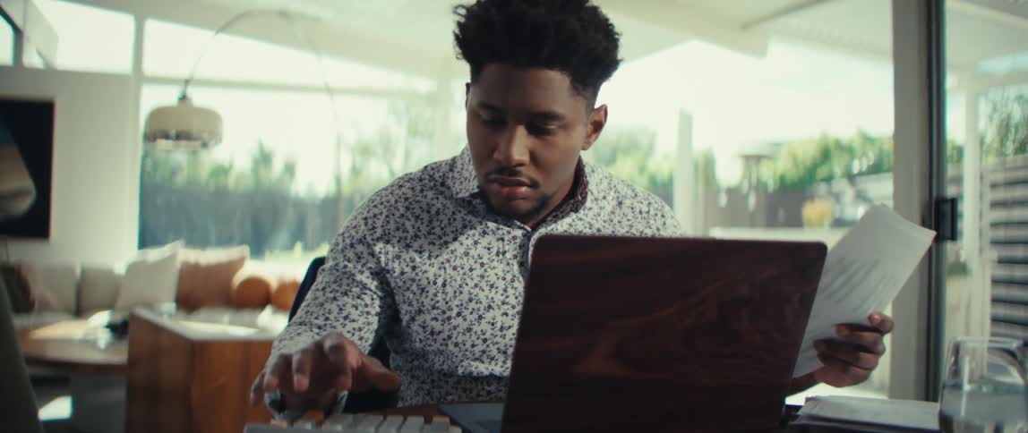 A young man sits at a wooden table in a bright room while looking at papers and typing on a laptop