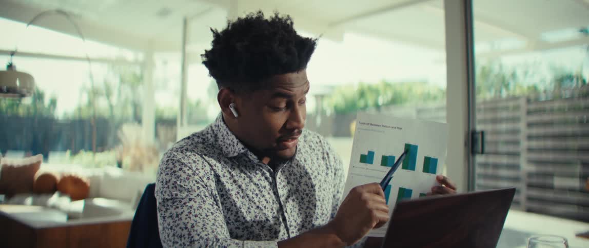 Young adult black man wearing a white dress shirt pointing at graphs on a piece of paper while talking in front of a laptop computer in a living room indoors during day time