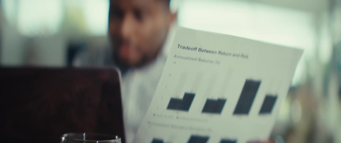 A man in a patterned shirt focuses on a laptop while holding a document titled tradeoff between return and risk that features a bar graphhe appears deep in thought looking back and forth between the physical paper and the computer screen in front of h